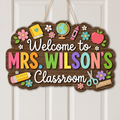A decorative wooden hanging sign that reads “Welcome to Mrs. Wilson’s Classroom,” decorated with colorful school-themed illustrations including flowers, a book, globe, crayon, apple, pencil, scissors, and ruler, shown hanging on a white paneled door.