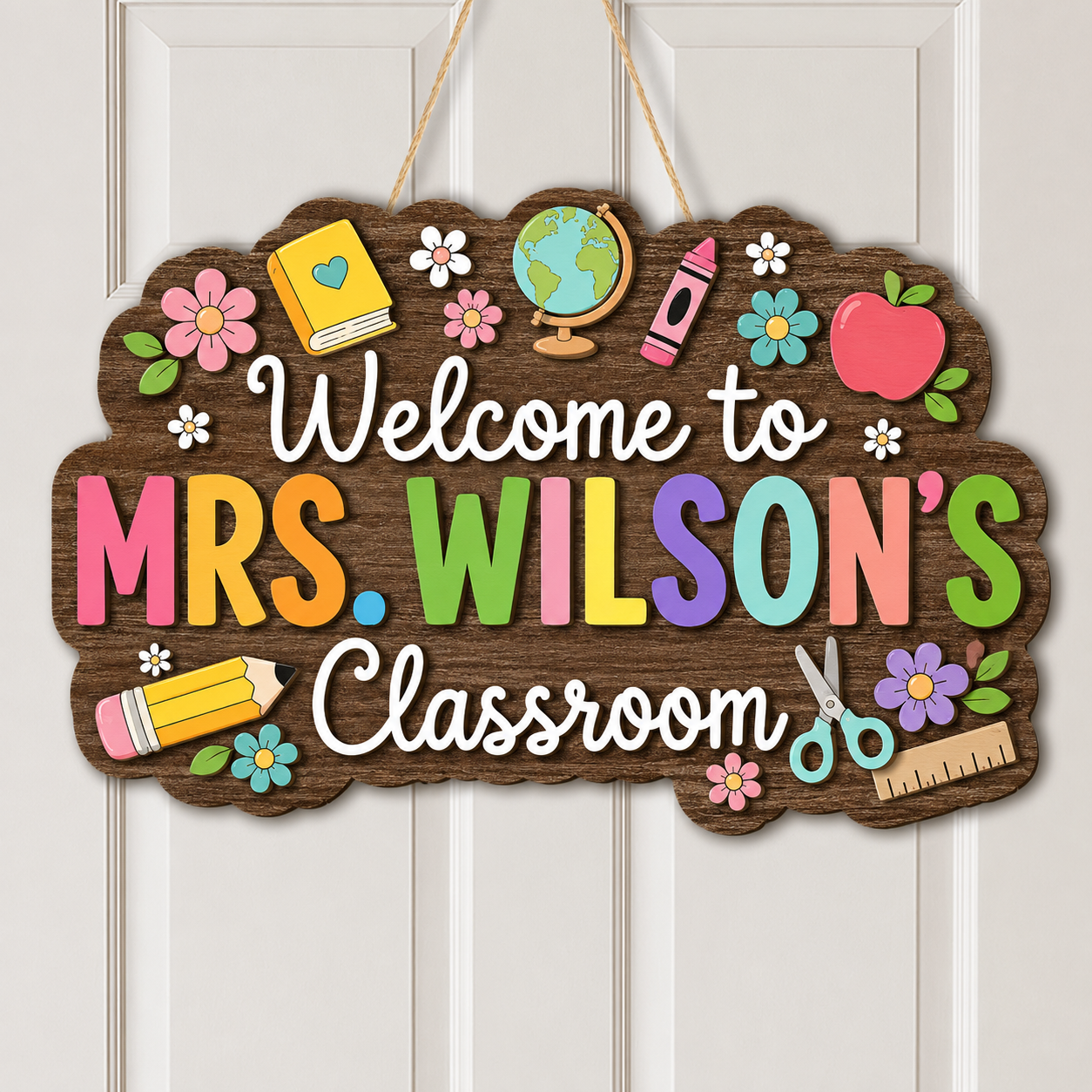 A decorative wooden hanging sign that reads “Welcome to Mrs. Wilson’s Classroom,” decorated with colorful school-themed illustrations including flowers, a book, globe, crayon, apple, pencil, scissors, and ruler, shown hanging on a white paneled door.