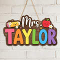 A personalized wooden hanging sign for a teacher, featuring the text 'Mrs. TAYLOR' in colorful letters on a brown wood background, with a smiling yellow pencil, red apple, and white flowers, hanging against a white brick wall.