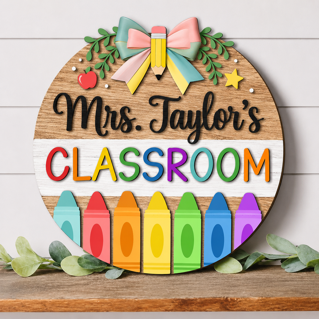 A round wooden classroom sign with the text 'Mrs. Taylor's CLASSROOM' in black and colorful lettering, decorated with a pastel bow, green leaves, a yellow pencil, stars, and a row of colorful crayons along the bottom.