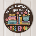 Personalized round wooden classroom door sign hanging on a white paneled door. The sign features a dark brown wood background with colorful decorative books, flowers, and the text 'IN THIS CLASSROOM you are MRS.