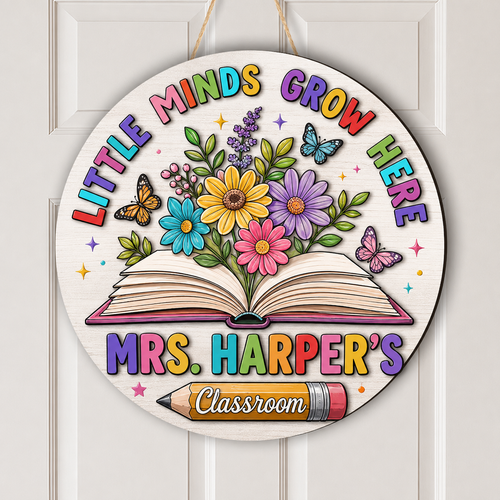 Round personalized wooden classroom door hanging sign with colorful text reading 'LITTLE MINDS GROW HERE' and 'MRS. HARPER'S Classroom' around an open book with flowers and butterflies, shown hanging on a white paneled door.