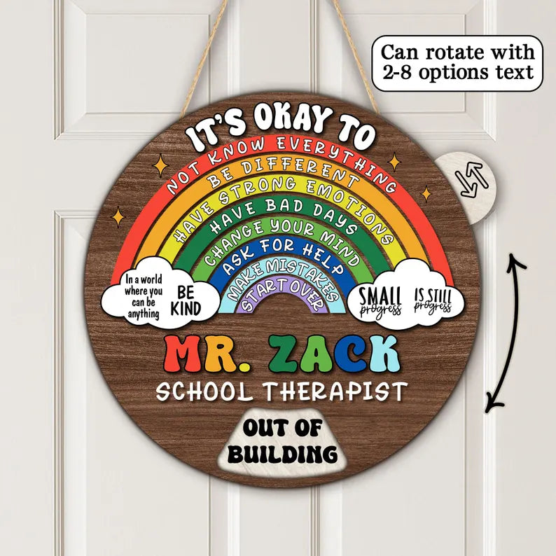 A round wooden hanging sign with a rainbow graphic and motivational text, shown on a white paneled door. The sign reads 'IT'S OKAY TO NOT KNOW EVERYTHING BE DIFFERENT HAVE BAD DAYS CHANGE YOUR MIND ASK FOR HELP WARMSTROUNDS MR.