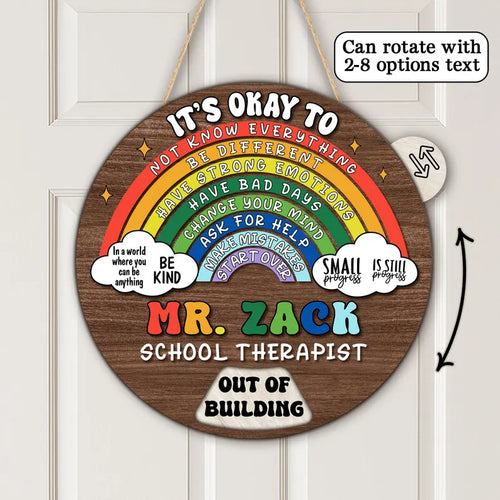 A round wooden hanging sign with a rainbow graphic and motivational text, shown on a white paneled door. The sign reads 'IT'S OKAY TO NOT KNOW EVERYTHING BE DIFFERENT HAVE BAD DAYS CHANGE YOUR MIND ASK FOR HELP WARMSTROUNDS MR.