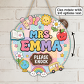 A round decorative hanging sign with the words “MRS. EMMA” in colorful letters and “PLEASE KNOCK” on a brown plaque, decorated with cute school-themed illustrations including a butterfly, sun, apple, pencil, book, globe, rainbow, clouds, and a yellow bus.