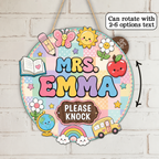A round decorative hanging sign with the words “MRS. EMMA” in colorful letters and “PLEASE KNOCK” on a brown plaque, decorated with cute school-themed illustrations including a butterfly, sun, apple, pencil, book, globe, rainbow, clouds, and a yellow bus.